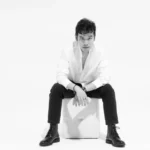 Jeffrey Chan seated on a white cube, wearing a white open-collar shirt, black trousers, and black lace-up boots, in a black and white studio portrait