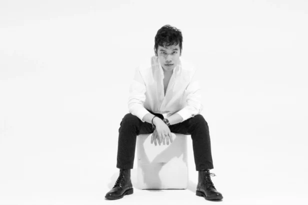 Jeffrey Chan seated on a white cube, wearing a white open-collar shirt, black trousers, and black lace-up boots, in a black and white studio portrait