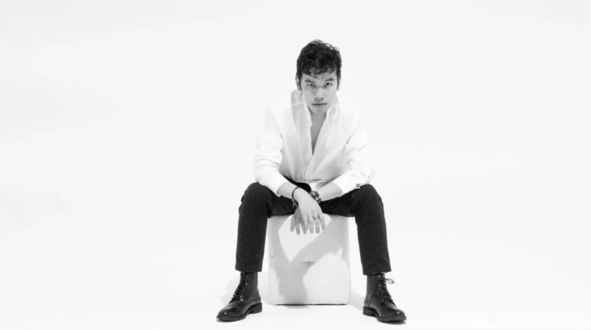 Jeffrey Chan seated on a white cube, wearing a white open-collar shirt, black trousers, and black lace-up boots, in a black and white studio portrait