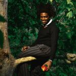 Kodak Black sitting on a tree branch in a lush green forest, holding an apple, with a snake resting on the branch beside him