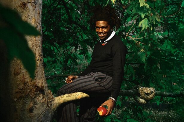 Kodak Black sitting on a tree branch in a lush green forest, holding an apple, with a snake resting on the branch beside him