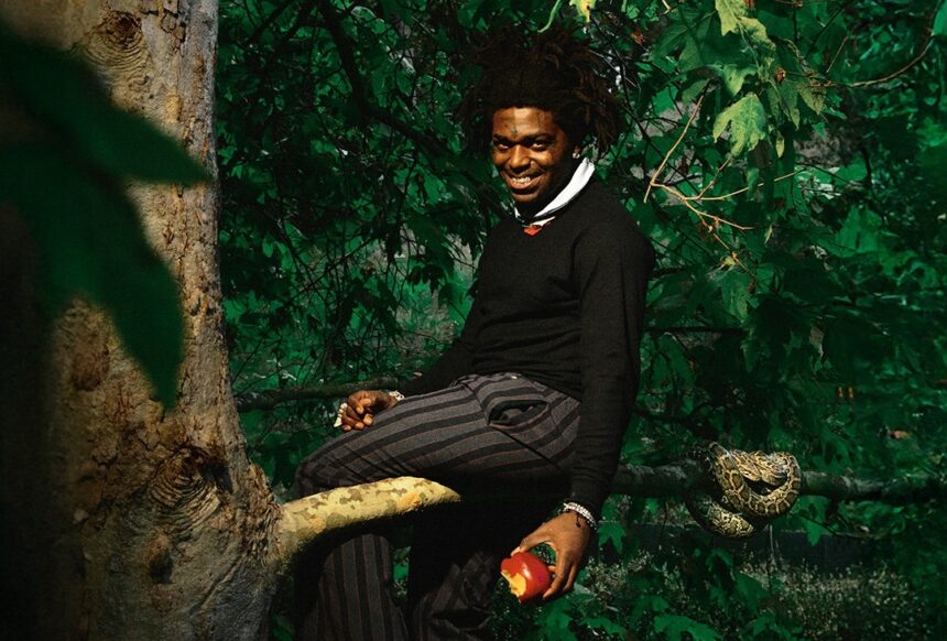 Kodak Black sitting on a tree branch in a lush green forest, holding an apple, with a snake resting on the branch beside him