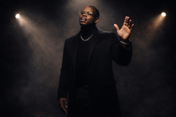 Mojo Marc performs on a dimly lit stage, dressed in an elegant black outfit with a gold chain and glasses, singing passionately under dramatic spotlights as soft haze fills the background.