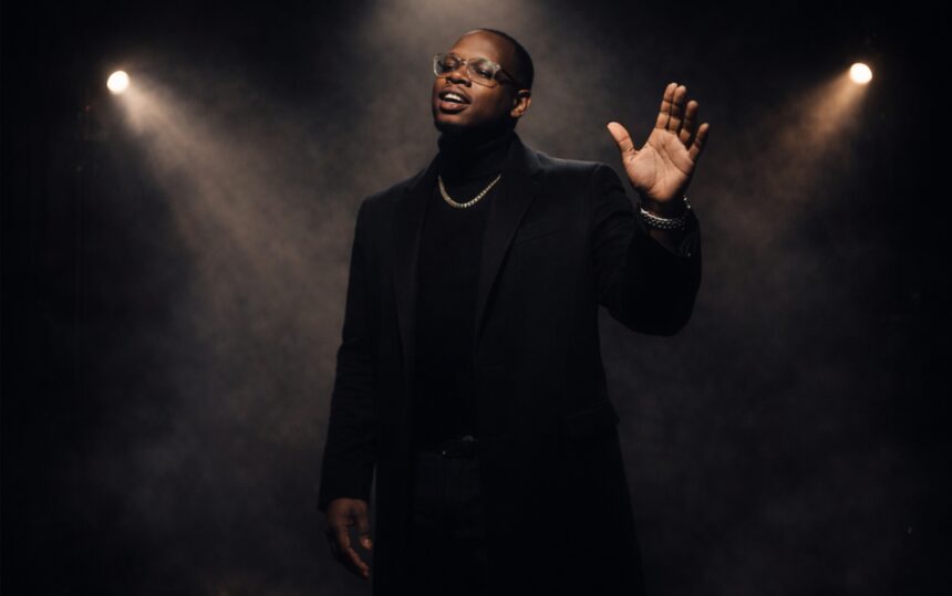 Mojo Marc performs on a dimly lit stage, dressed in an elegant black outfit with a gold chain and glasses, singing passionately under dramatic spotlights as soft haze fills the background.