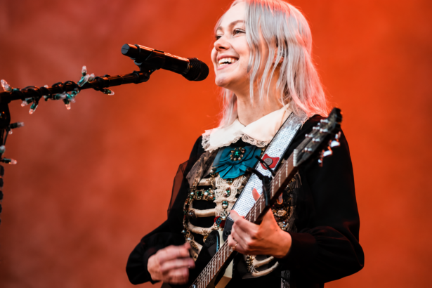 Phoebe Bridgers performing live on stage during The Lost Tour 2026, smiling while playing guitar in a black embroidered dress