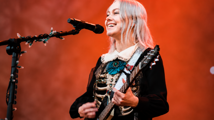 Phoebe Bridgers performing live on stage during The Lost Tour 2026, smiling while playing guitar in a black embroidered dress
