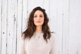 Sara Bareilles standing in front of a weathered white wooden wall, wearing a light knit sweater and looking calmly at the camera in natural daylight.