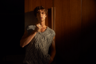 Shawn Mendes in a gray knit sweater, lit by warm golden light against dark wood panels.