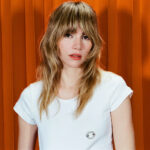 Suki Waterhouse in a white t-shirt against an orange curtain backdrop.