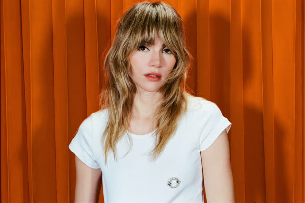 Suki Waterhouse in a white t-shirt against an orange curtain backdrop.