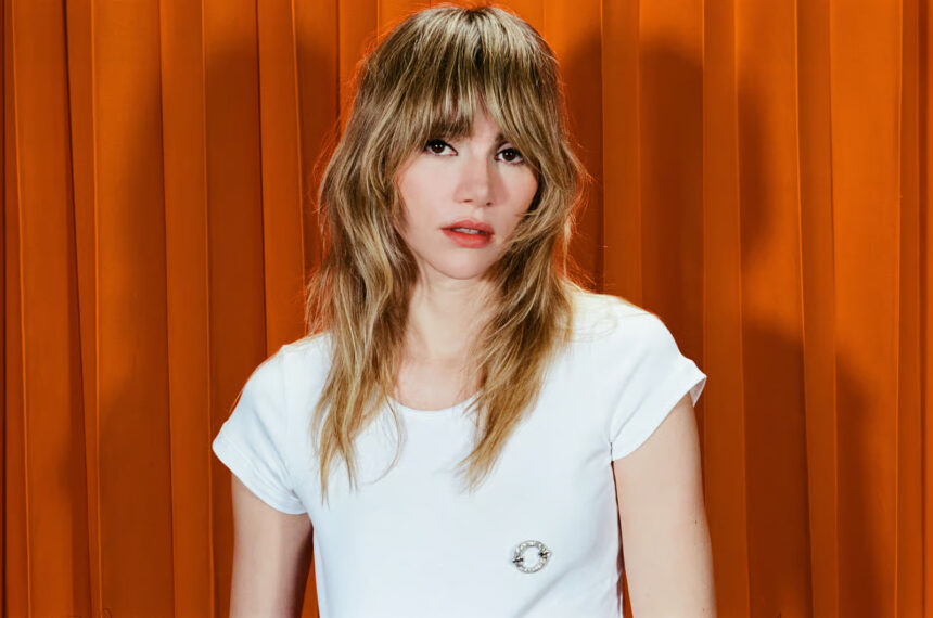 Suki Waterhouse in a white t-shirt against an orange curtain backdrop.