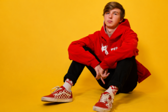 Electronic music producer Whethan in a red hoodie and checkered Vans sneakers posed against a yellow studio background ahead of his 2026 WAREHOUSE.WAVS Tour across North America