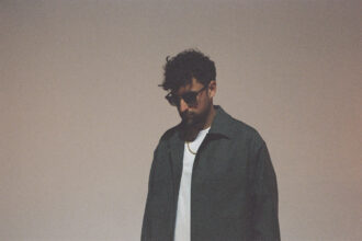 Wuki, a man with curly dark hair and sunglasses, wearing a dark jacket over a white t-shirt and a gold chain necklace, looking downward against a soft beige background
