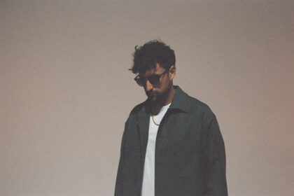 Wuki, a man with curly dark hair and sunglasses, wearing a dark jacket over a white t-shirt and a gold chain necklace, looking downward against a soft beige background