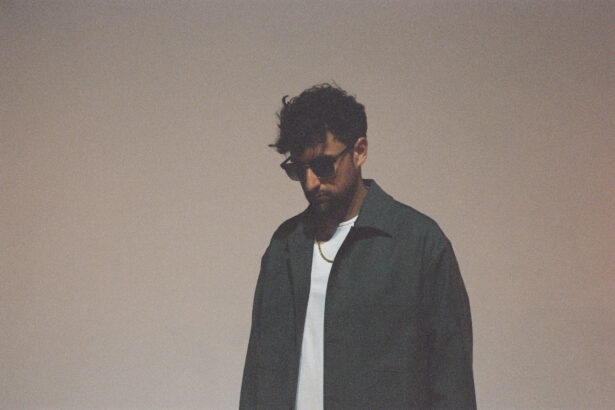 Wuki, a man with curly dark hair and sunglasses, wearing a dark jacket over a white t-shirt and a gold chain necklace, looking downward against a soft beige background