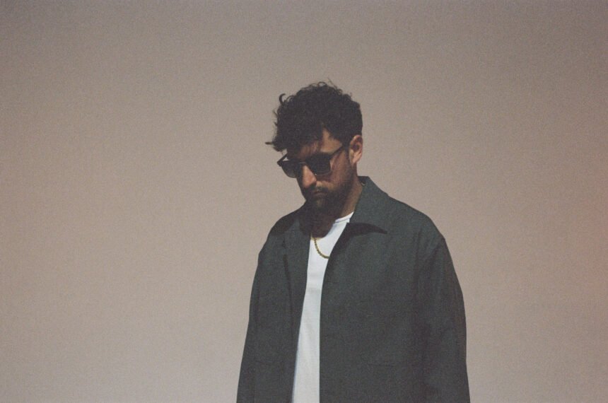 Wuki, a man with curly dark hair and sunglasses, wearing a dark jacket over a white t-shirt and a gold chain necklace, looking downward against a soft beige background