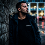 Liverpool trance producer AMOS & RIOT NIGHT leans against a stone wall on a rainy city street ahead of new single Pieces.