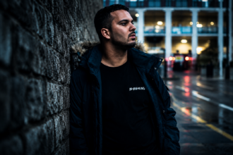Liverpool trance producer AMOS & RIOT NIGHT leans against a stone wall on a rainy city street ahead of new single Pieces.