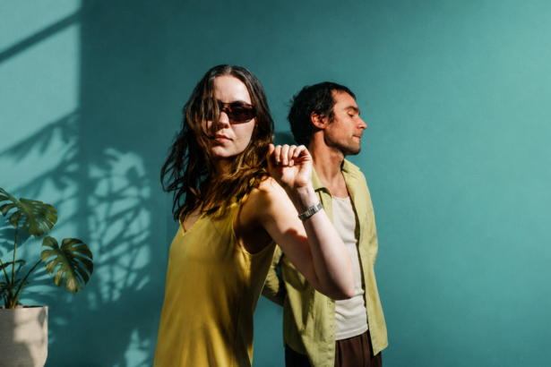 Sam and Louise Sullivan pose back to back against a teal wall in warm sunlight for their 'Love and Devotion' press photo