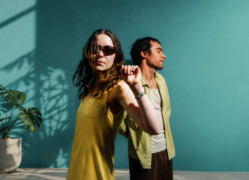 Sam and Louise Sullivan pose back to back against a teal wall in warm sunlight for their 'Love and Devotion' press photo