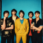 The Strokes pose against a blue backdrop, frontman Julian Casablancas centered in a yellow suit