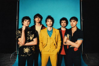The Strokes pose against a blue backdrop, frontman Julian Casablancas centered in a yellow suit