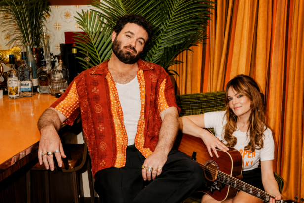 Angus & Julia Stone pose in a retro bar setting with orange curtains, Julia holding an acoustic guitar, in a press photo for their album 'Karaoke Bar.'