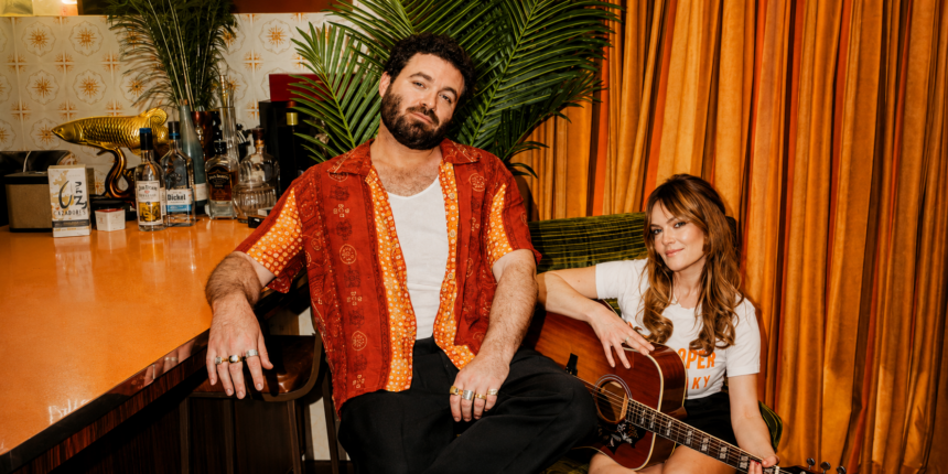 Angus & Julia Stone pose in a retro bar setting with orange curtains, Julia holding an acoustic guitar, in a press photo for their album 'Karaoke Bar.'