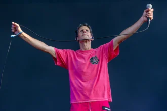 Mike D of the Beastie Boys performs live in a pink shirt, arms outstretched holding a microphone and cable against a dark stage