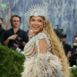 Beyoncé smiling over her shoulder at the Met Gala in a crystal headpiece and feathered gown, with photographers behind her