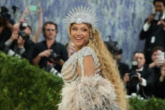 Beyoncé smiling over her shoulder at the Met Gala in a crystal headpiece and feathered gown, with photographers behind her