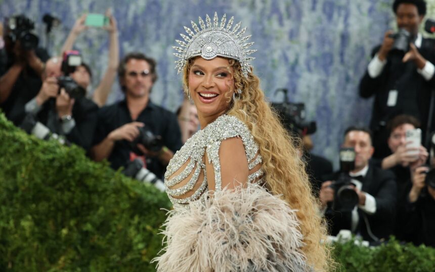 Beyoncé smiling over her shoulder at the Met Gala in a crystal headpiece and feathered gown, with photographers behind her