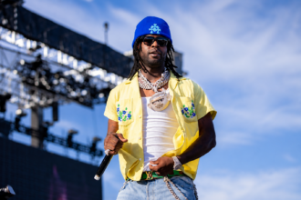 Chief Keef performs on stage at Rolling Loud 2024, wearing a blue beanie, sunglasses, chain necklaces, and a yellow floral shirt over a white tank top while holding a microphone