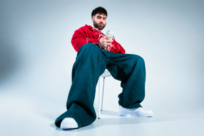 Fat Papi sitting on a white chair in a minimalist studio, wearing a red hoodie