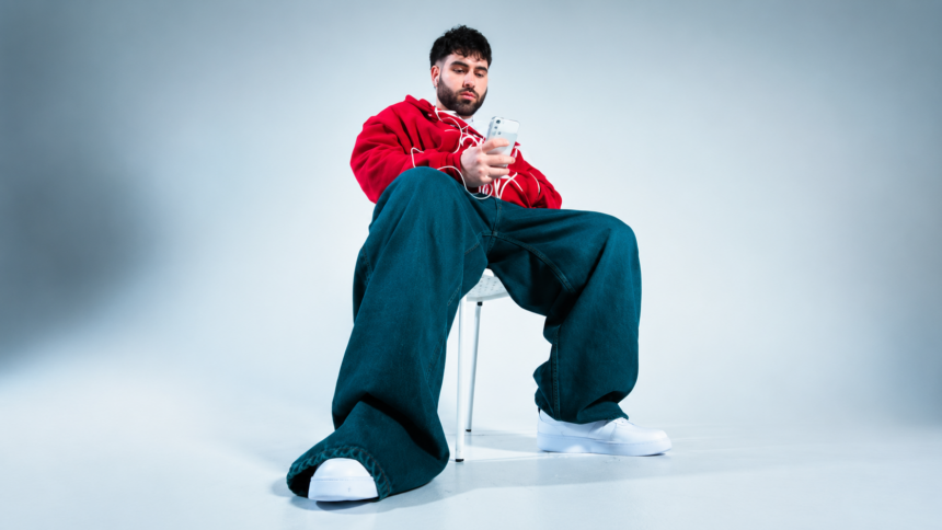 Fat Papi sitting on a white chair in a minimalist studio, wearing a red hoodie