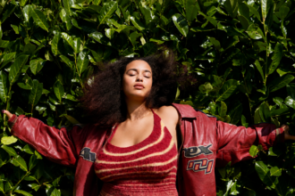 Jorja Smith stands with her arms outstretched against a lush wall of green leaves