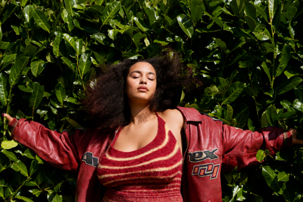 Jorja Smith stands with her arms outstretched against a lush wall of green leaves