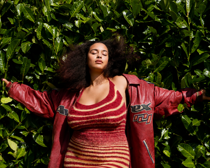 Jorja Smith stands with her arms outstretched against a lush wall of green leaves