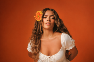 Karol G, wearing a white dress with a flower in her hair against an orange background, in a press photo for 'No Me Arrepiento de Sentir Tanto.'