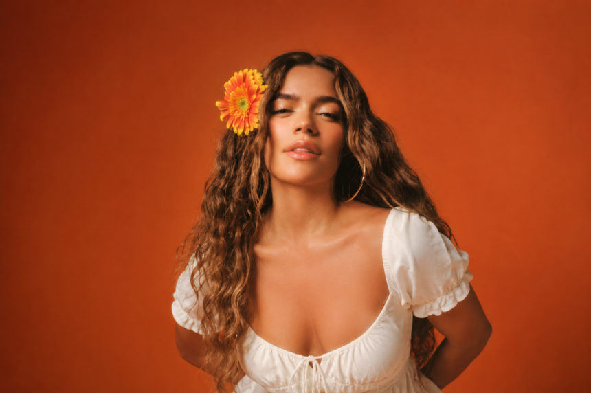 Karol G, wearing a white dress with a flower in her hair against an orange background, in a press photo for 'No Me Arrepiento de Sentir Tanto.'