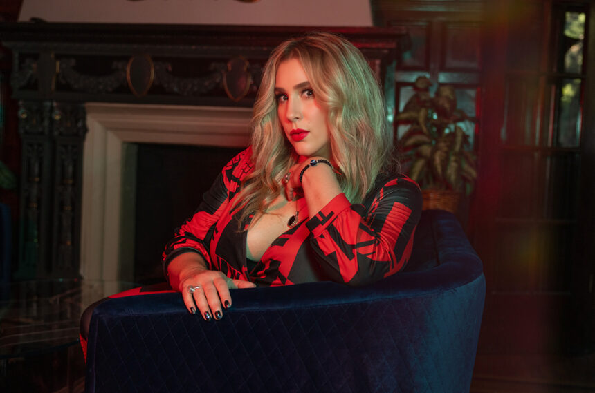 KiKi Holli poses in a red and black patterned dress on a blue velvet chair in a moody, vintage room, in a press photo for the EP 'Something About You.'