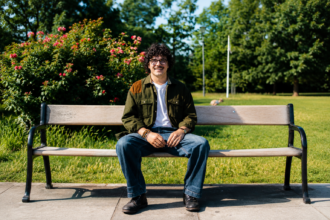 Indie-folk musician Luca Firth smiles while sitting on a park bench in a green jacket and glasses, photographed around the release of 'Big Eagle Hunting in the Wind.'