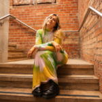 Lucy Robinson sits on brick-lined stairs in a colourful pastel dress ahead of her new alt-pop single Let Down.