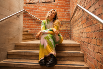 Lucy Robinson sits on brick-lined stairs in a colourful pastel dress ahead of her new alt-pop single Let Down.
