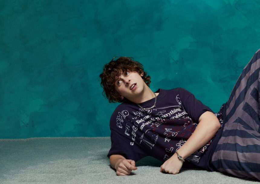 NOAHFINNCE Noah Adams lying on a studio floor wearing a graphic-print t-shirt and layered jewelry, promotional image for his Mr Overwhelm UK Tour in October 2026