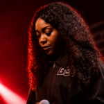 Chicago rapper Noname performs on stage under red lighting, holding a microphone and looking down