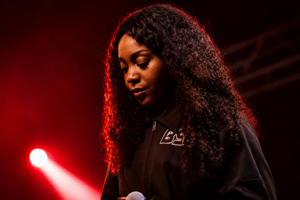 Chicago rapper Noname performs on stage under red lighting, holding a microphone and looking down