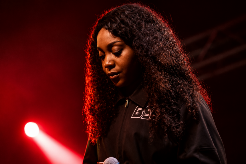 Chicago rapper Noname performs on stage under red lighting, holding a microphone and looking down