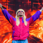 Oliver Tree performs onstage with arms raised, wearing a blond mullet wig and pink and purple windbreaker