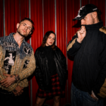 Brighton alt-punk trio SNAYX pose against a red corrugated wall in a press photo for 'Nice Day 4 It'